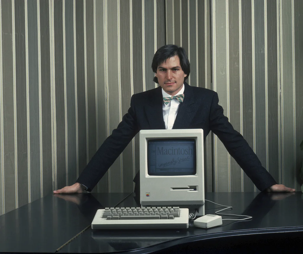 Steve Jobs präsentiert mit einer Fliege um den Hals den ersten Apple Macintosh. 